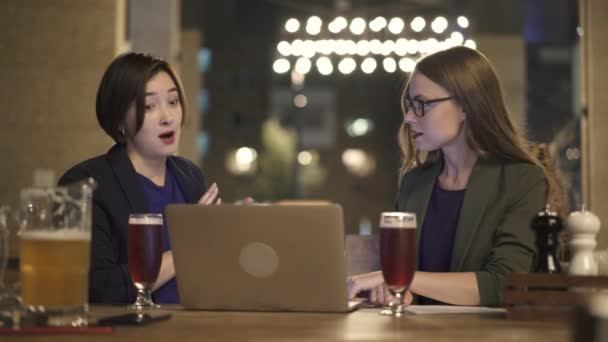 Dos mujeres jóvenes con portátil hablando en la cafetería al aire libre por la noche — Vídeo de stock