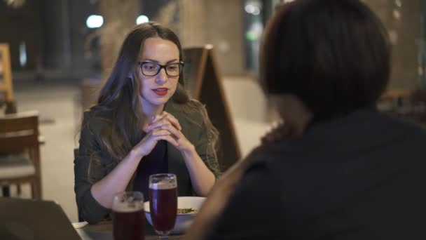 Привлекательная молодая женщина в очках разговаривает со своей подругой в ночном кафе — стоковое видео