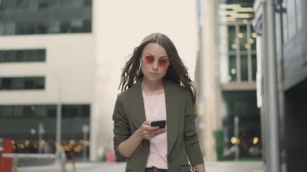 Спокойная женщина в красных солнцезащитных очках ходит по улице и смотрит на экран смартфона — стоковое видео