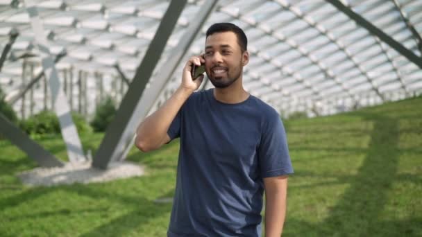 Усміхнений молодий чоловік розмовляє по телефону, стоячи на зеленому газоні — стокове відео