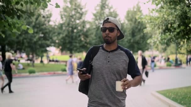 Cheerful young man in baseball cap walking in the street and drinking coffee — Stock Video