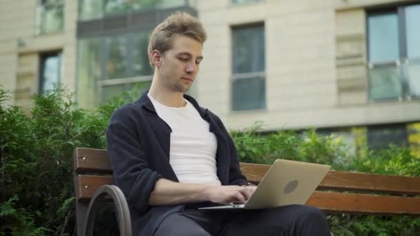 Νεαρός άνδρας πληκτρολογώντας σε φορητό υπολογιστή εκτός και ελέγχοντας το ρολόι του — Αρχείο Βίντεο