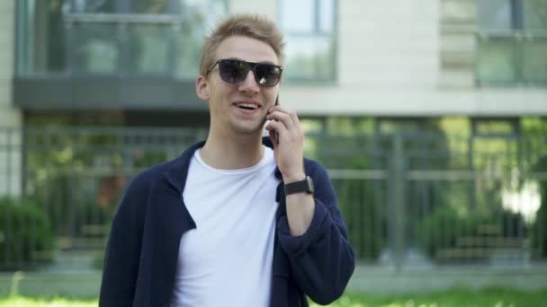Человек в солнечных очках смеется, разговаривая по смартфону — стоковое видео