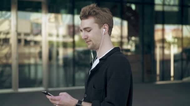 Vista lateral de un joven sonriente con smartphone y café escuchando música — Vídeo de stock