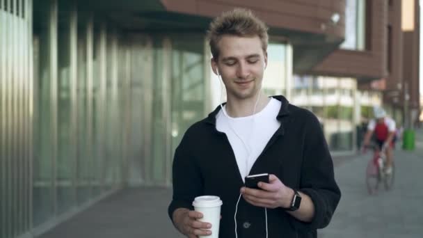 Framifrån leende ung man med smartphone och kaffe som lyssnar på musik — Stockvideo