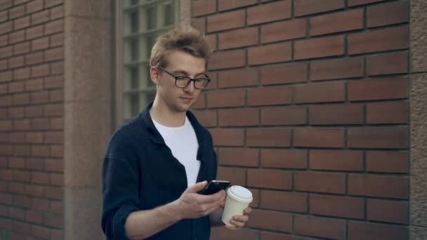 Człowiek w okularach, chodzenie w pobliżu budynku z cegły, picia kawy i surfowanie po Internecie — Wideo stockowe