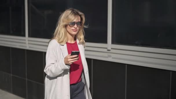 Веселая блондинка в солнечных очках ходит и смотрит на телефон — стоковое видео