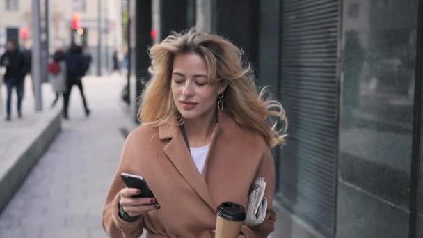 Atractiva mujer rubia con café caminando y usando su smartphone — Vídeos de Stock