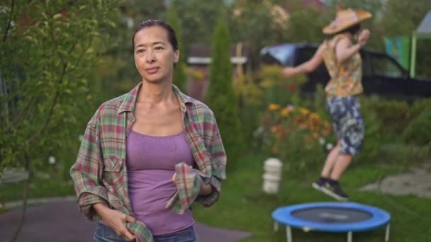 Mamma står i trädgården medan dottern hoppar på studsmatta — Stockvideo