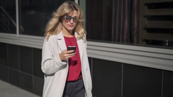 Улыбающаяся блондинка в солнечных очках ходит и смотрит на телефон — стоковое видео