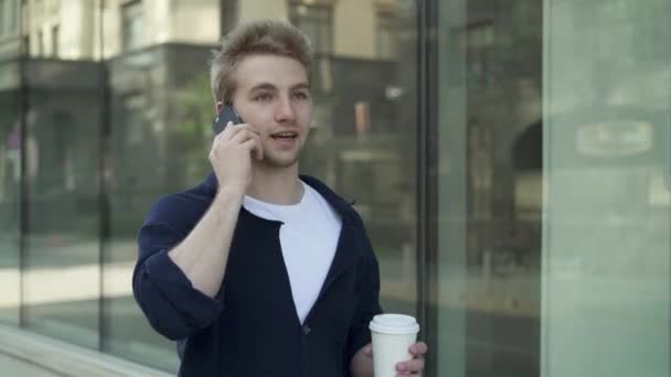 Jonge man met koffie wandelen langs bouwen en praten over de telefoon — Stockvideo