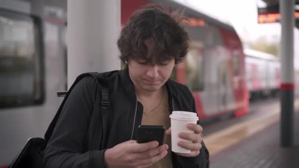 Joven con teléfono bebiendo café en la estación de tren — Vídeo de stock