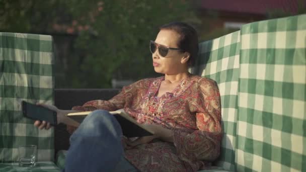 De mediana edad mujer asiática leyendo un libro y revisando su teléfono — Vídeos de Stock