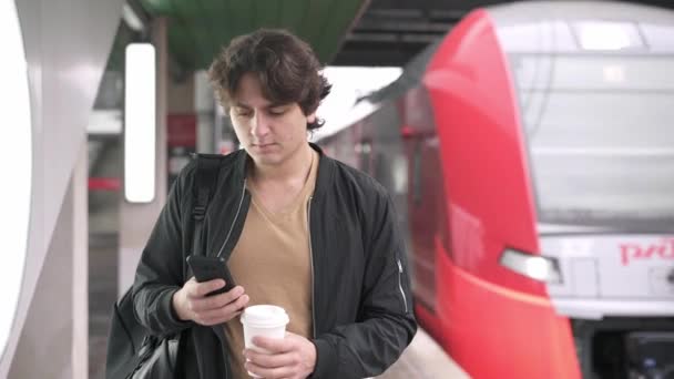 ハンサムな若い男コーヒーを飲みながら鉄道駅プラットフォームに沿って歩いて行くと Web サーフィン 鉄道が到着します ミディアム ショットのスローモーションを追跡 — ストック動画