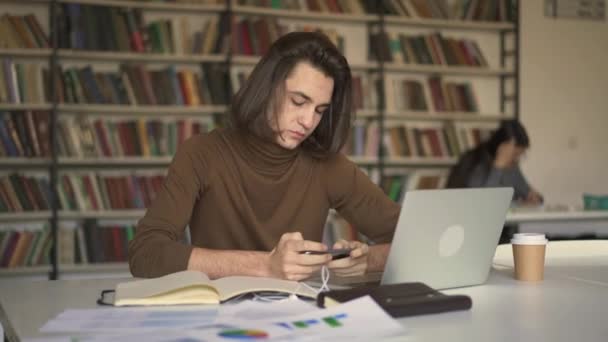 Joven con el pelo largo golpeando en un teléfono inteligente en la biblioteca — Vídeos de Stock