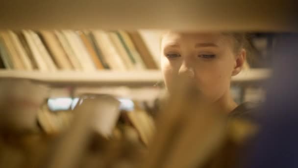 Ξανθιά κοπέλα επιλέγοντας ένα βιβλίο στο ράφι της βιβλιοθήκης — Αρχείο Βίντεο