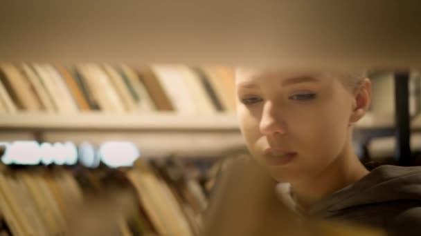 Blond meisje het kiezen van een boek op de plank in de bibliotheek — Stockvideo