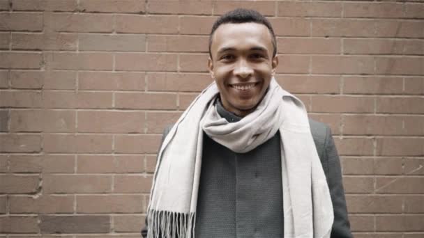 Handsome African American man wearing a coat and a scarf is smiling toward camera. Handheld slow motion portrait shot. Brick wall on background — Stock Video