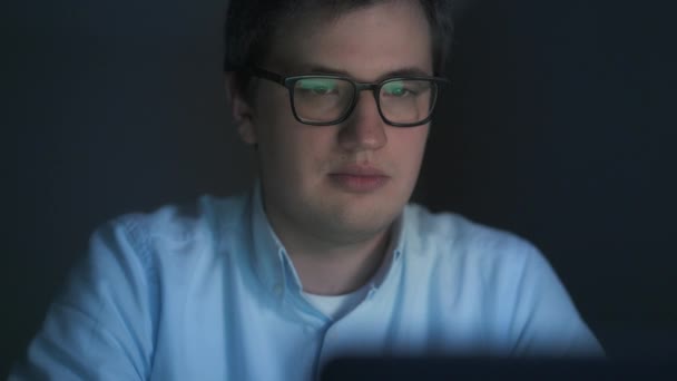 Νεαρός άνδρας με γυαλιά που εργάζονται για το laptop το βράδυ σε ένα σκοτεινό δωμάτιο — Αρχείο Βίντεο