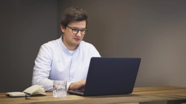 Erwachsener Mann mit Brille sitzt am Tisch und tippt lächelnd auf Laptop — Stockvideo