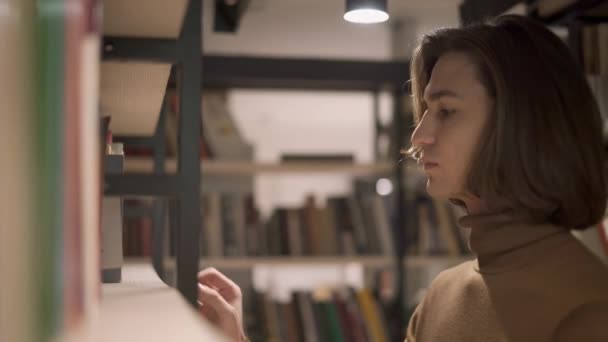 Молодой человек берет книгу на полке в библиотеке — стоковое видео