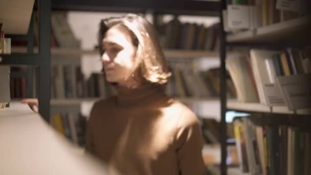 Молодой человек выбирает книгу на полке в библиотеке — стоковое видео