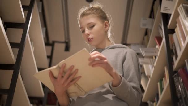 Mladá dívka obrací stránky knihy v knihovně, stojící mezi regály — Stock video