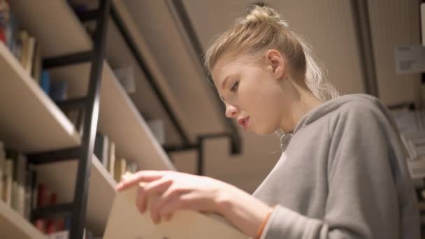 Ung flicka vänder sidor i en bok i biblioteket står mellan bokhyllor — Stockvideo