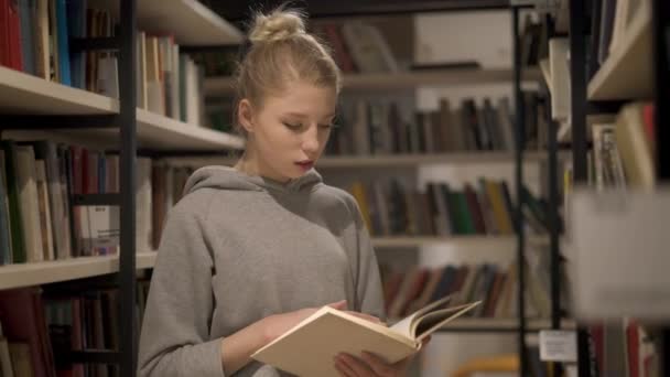 Ung blond tjej läser en bok stående mellan bokhyllorna i biblioteket — Stockvideo