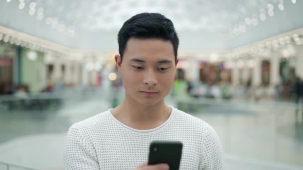 Портрет азиатского мужчины, прокручивающего телефон на фоне большого торгового центра — стоковое видео