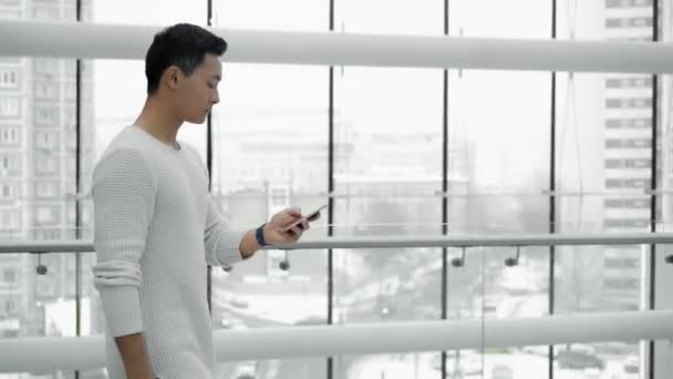 窓の背景に携帯電話で歩くアジアの男性のミドルショットプロファイル — ストック動画