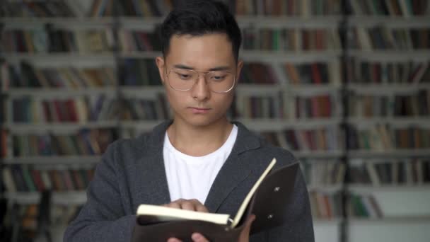 Молодой человек пролистывает блокнот на фоне библиотечных полок — стоковое видео