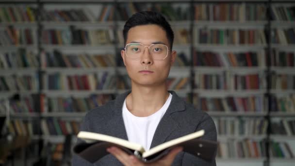 Ein asiatischer Mann mit einer Blocknote steht in der Bibliothek und blickt geradeaus — Stockvideo