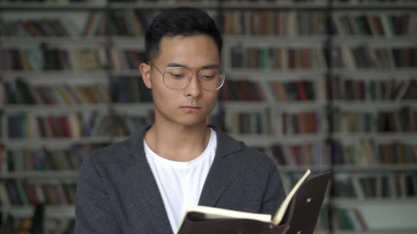Молодой человек читает блокнот, размышляя на фоне книжных полок — стоковое видео