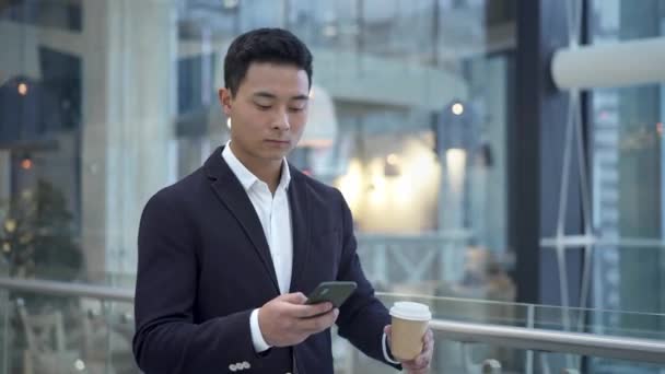 Азіатський молодий бізнесмен п'є кавові повідомлення по телефону в бізнес-центрі — стокове відео