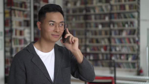 Азиатский юноша разговаривает по телефону на фоне книжных полок — стоковое видео