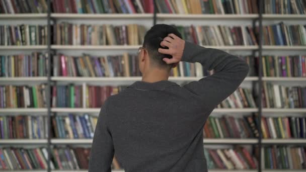 Цветной человек выбирает книгу на книжной полке, чешет голову — стоковое видео