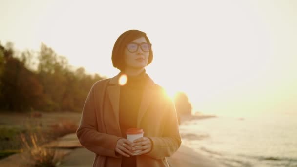 Mouvement lent de la femme asiatique en manteau et lunettes de vue marchant sur la plage au coucher du soleil — Video