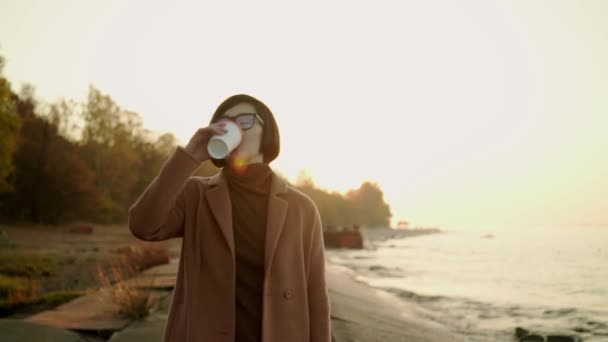 Mouvement lent de la femme asiatique en manteau et lunettes de vue marchant sur la plage au coucher du soleil — Video