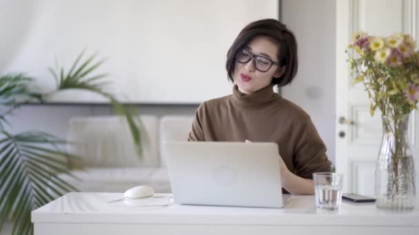 Азиатка в очках, имеющая видео чат в ноутбуке в домашнем офисе — стоковое видео