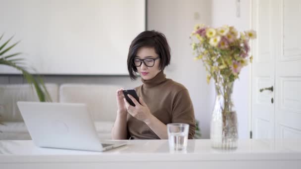 Азиатская женщина прокручивает телефон сидя в белом домашнем офисе — стоковое видео