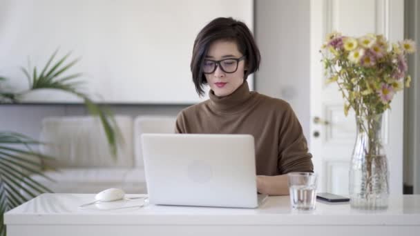 Asiatin mit Brille schreibt in Laptop im weißen Home Office — Stockvideo