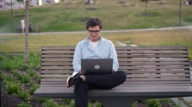 Genç adam parkta dizüstü bilgisayar kullanıyor bulanık çim arka planda bankta oturuyor.