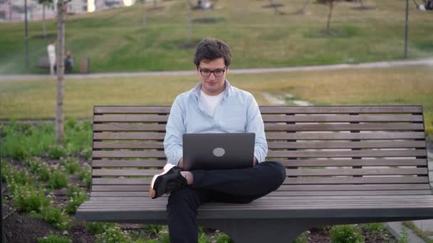 若いです男使用してノートパソコンで公園に座っている上のベンチにぼやけた草の背景 — ストック動画
