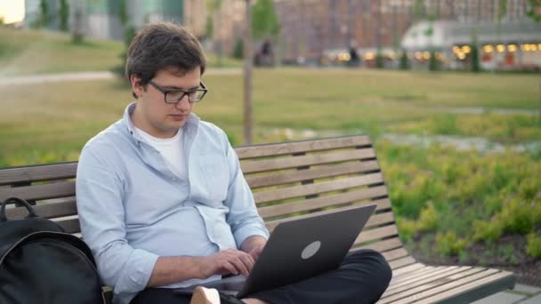在公园里的笔记本电脑上打字的年轻人坐在模糊的公园背景上的长椅上 — 图库视频影像