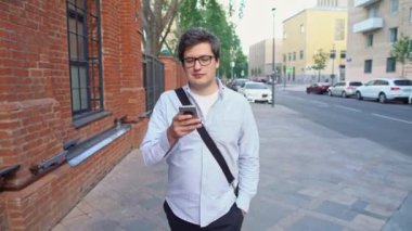 Gözlüklü ve mavi gömlekli bir adamın caddede telefonla konuşurken orta pozu.