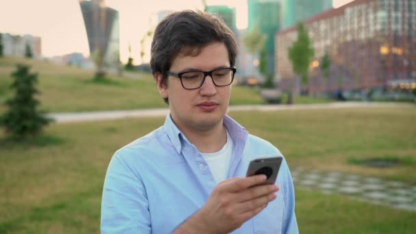 Портретний панорамний знімок дорослого чоловіка, який використовує мобільний телефон на фоні паркового газону — стокове відео