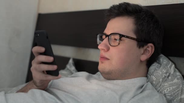 Jeune homme en lunettes avec un téléphone couché au lit — Video