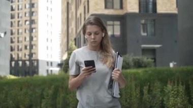 Genç bir kadın dizüstü bilgisayarla telefonda yazıyor, yol boyunca yürüyor.