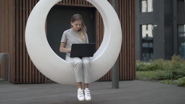 Jeune femme avec ordinateur portable en swing futuriste blanc dans la cour à l'extérieur — Video